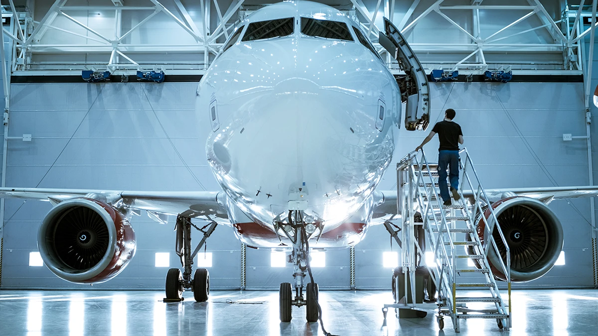 Aircraft in a hangar with a technician inspecting the nose on a maintenance platform.